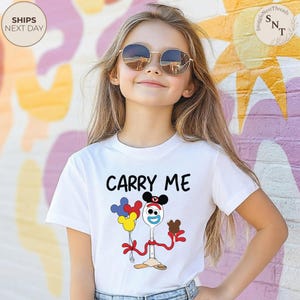 May include: White t-shirt with the text "CARRY ME" above a cartoon character holding balloons and a chocolate treat. The character is a spoon with a face and arms. The shirt is worn by a young person wearing sunglasses.