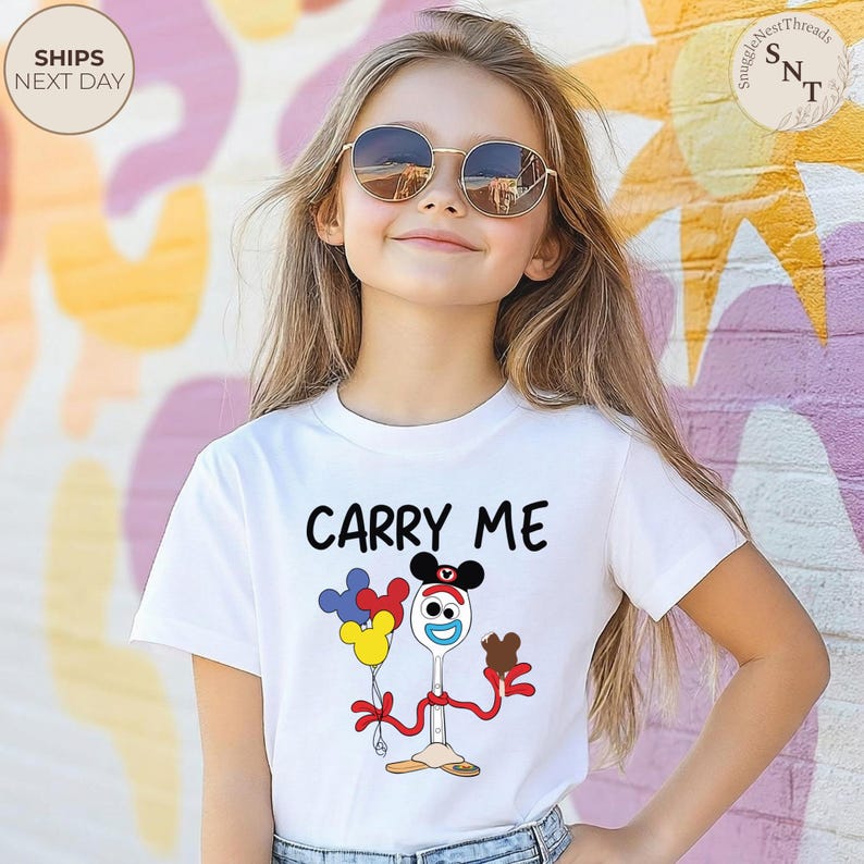 May include: White t-shirt with the text "CARRY ME" above a cartoon character holding balloons and a chocolate treat. The character is a spoon with a face and arms. The shirt is worn by a young person wearing sunglasses.