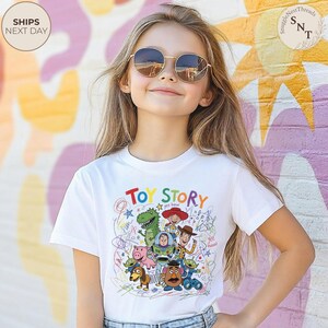 May include: White t-shirt featuring a colorful "Toy Story" graphic with various characters and the text "Toy Story" in rainbow colors. The design includes Woody, Buzz Lightyear, and other characters from the movie.