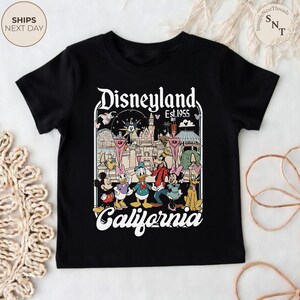 May include: A black short-sleeve t-shirt featuring a graphic print of Disneyland California Est. 1955. The design includes a castle illustration and cartoon characters including Mickey Mouse, Minnie Mouse, Donald Duck, Daisy Duck, Goofy, and Pluto.