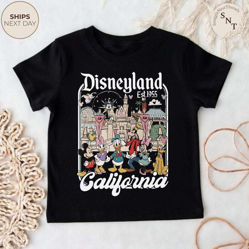 May include: A black short-sleeve t-shirt featuring a graphic print of Disneyland California Est. 1955. The design includes a castle illustration and cartoon characters including Mickey Mouse, Minnie Mouse, Donald Duck, Daisy Duck, Goofy, and Pluto.