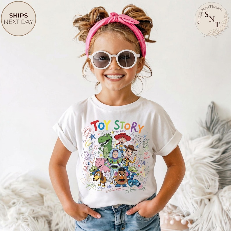 May include: A white t-shirt featuring a colorful graphic of Toy Story characters and the words "Toy Story" in a playful font. The design includes Woody, Buzz Lightyear, and other characters. The shirt is worn by a young person.