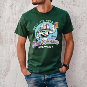 Puede incluir: Camiseta verde oscuro con una ilustraci&oacute;n de dibujos animados de Buzz Lightyear sosteniendo una jarra de cerveza. El dise&ntilde;o incluye el texto "Buzz Lite Beer" y "Star Command Brewery" con "Best Quality Since 1995".