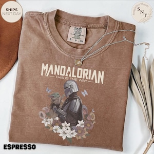 Puede incluir: Una camiseta marr&oacute;n de manga corta con un gr&aacute;fico del Mandaloriano y Grogu rodeados de flores y mariposas. El texto dice MANDALORIAN THIS IS THE WAY. La camiseta tiene cuello redondo y una etiqueta de la marca Comfort Colors en el interior.