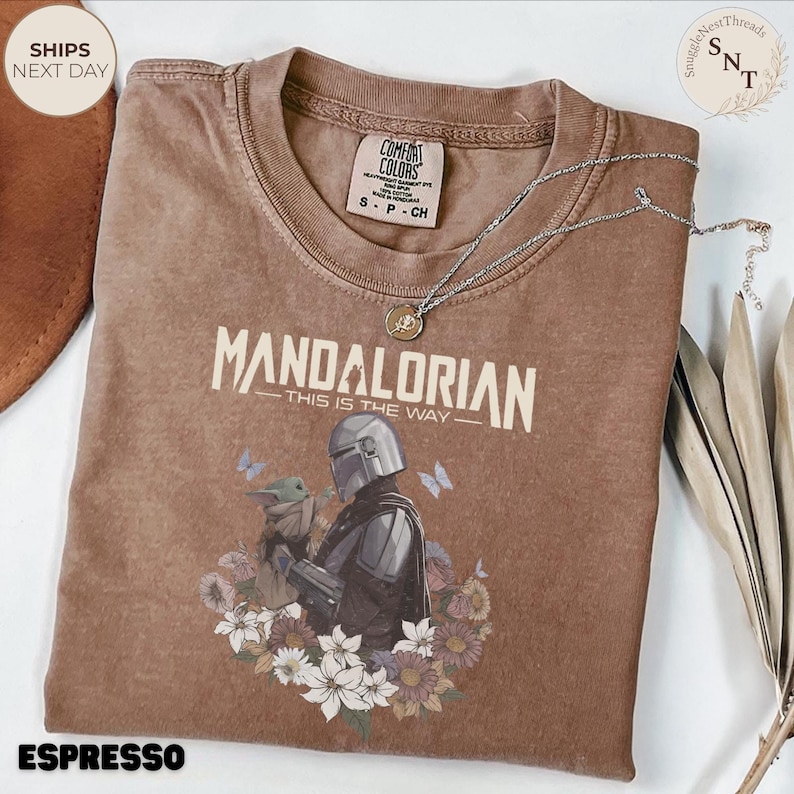 Puede incluir: Una camiseta marr&oacute;n de manga corta con un gr&aacute;fico del Mandaloriano y Grogu rodeados de flores y mariposas. El texto dice MANDALORIAN THIS IS THE WAY. La camiseta tiene cuello redondo y una etiqueta de la marca Comfort Colors en el interior.