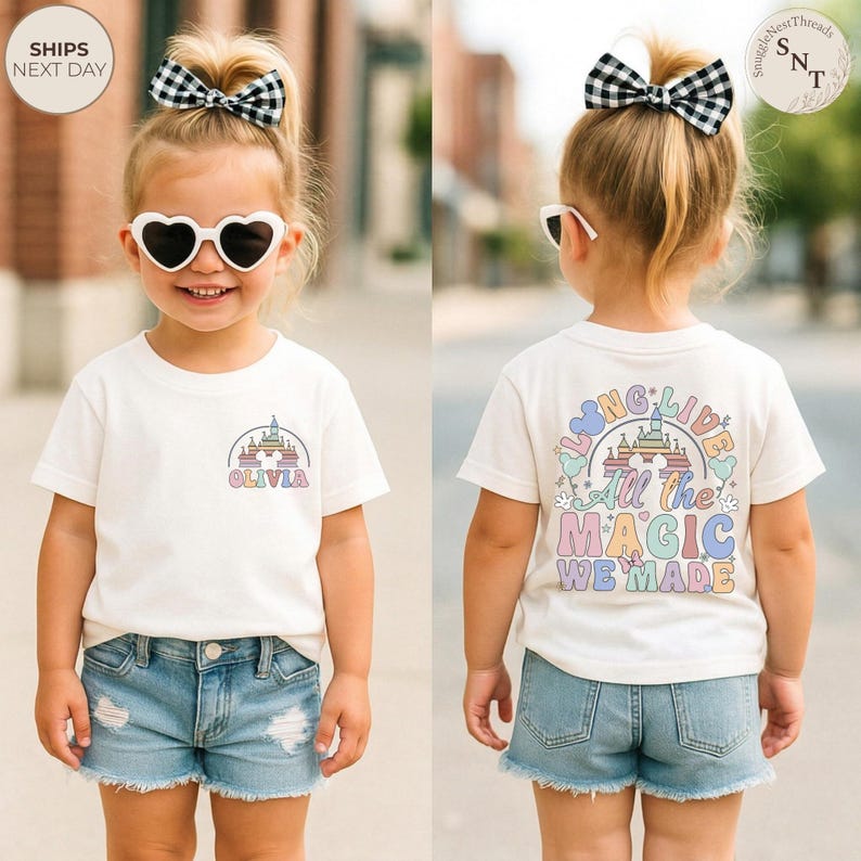 May include: A cream-colored t-shirt with a colorful castle graphic and the name "OLIVIA" printed on the front. The back of the shirt features the text "LIVING LIVE All the MAGIC WE MADE" in pastel colors. The child is wearing denim shorts and heart-shaped sunglasses.