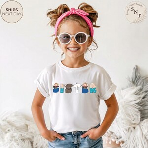 K&ouml;nnte beinhalten: Ein wei&szlig;es T-Shirt mit dem Wort "SISTER" auf der Vorderseite in bunten, cartoonartigen Buchstaben. Die Buchstaben zeigen Illustrationen von Figuren. Das Kind tr&auml;gt eine Sonnenbrille und ein rosa Stirnband.