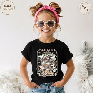 Pu&ograve; includere: Una maglietta nera con una grafica dei personaggi Disney, tra cui Topolino e Minnie, Paperino e Paperina, e Pippo, con la scritta "Animal Kingdom" e "Walt Disney World". La maglietta &egrave; indossata da un bambino.