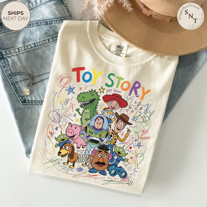 May include: Cream-colored t-shirt featuring a colorful graphic of Toy Story characters, including Woody, Buzz Lightyear, and Rex. The words "Toy Story" are written in a playful font, with "yee haw" included. The shirt is folded on top of blue jeans.
