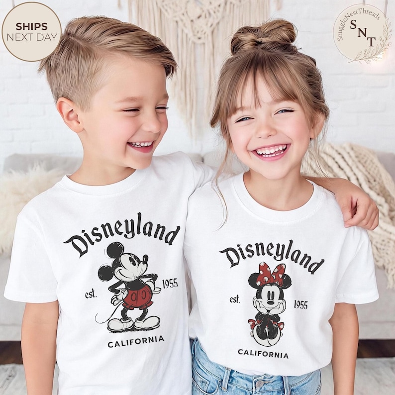 Pu&ograve; includere: Due bambini sorridenti indossano magliette bianche con una grafica di Disneyland. Le magliette presentano Topolino e Minnie, la scritta "Disneyland", "est. 1955" e "California".