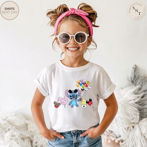 Pu&ograve; includere: T-shirt bianca per bambini con una grafica a cartoni animati colorata di un personaggio blu che tiene in mano palloncini, zucchero filato e coni gelato. La ragazza indossa occhiali da sole e una fascia rosa.