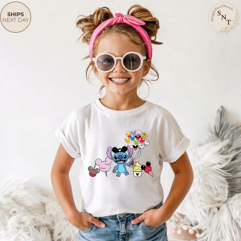 Pu&ograve; includere: T-shirt bianca per bambini con una grafica a cartoni animati colorata di un personaggio blu che tiene in mano palloncini, zucchero filato e coni gelato. La ragazza indossa occhiali da sole e una fascia rosa.