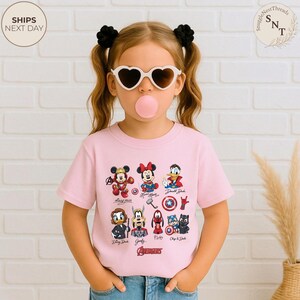 May include: A light pink t-shirt featuring cartoon characters including Mickey Mouse, Minnie Mouse, and Donald Duck. The word "AVENGERS" is printed at the bottom of the shirt. The child is wearing heart-shaped sunglasses and has black hair ties.