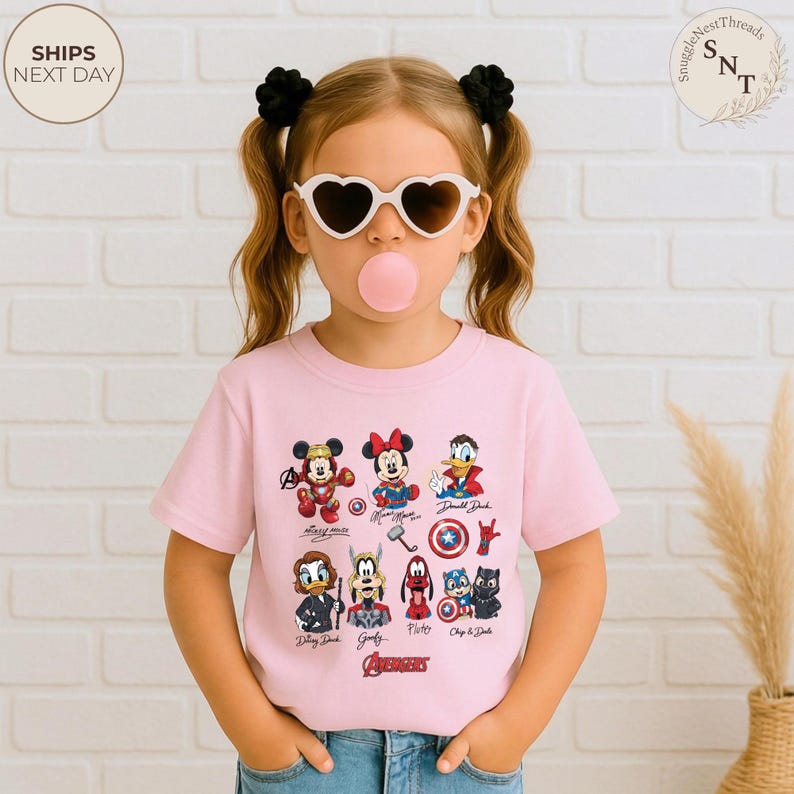 May include: A light pink t-shirt featuring cartoon characters including Mickey Mouse, Minnie Mouse, and Donald Duck. The word "AVENGERS" is printed at the bottom of the shirt. The child is wearing heart-shaped sunglasses and has black hair ties.