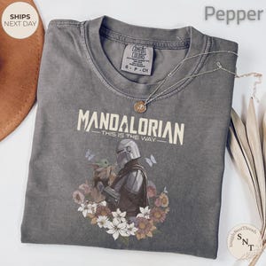 Puede incluir: Una camiseta de cuello redondo color pimienta con un gr&aacute;fico de The Mandalorian y Grogu rodeados de flores y mariposas. El texto dice "MANDALORIAN THIS IS THE WAY". Un collar de tono plateado con un peque&ntilde;o colgante descansa sobre la camiseta.