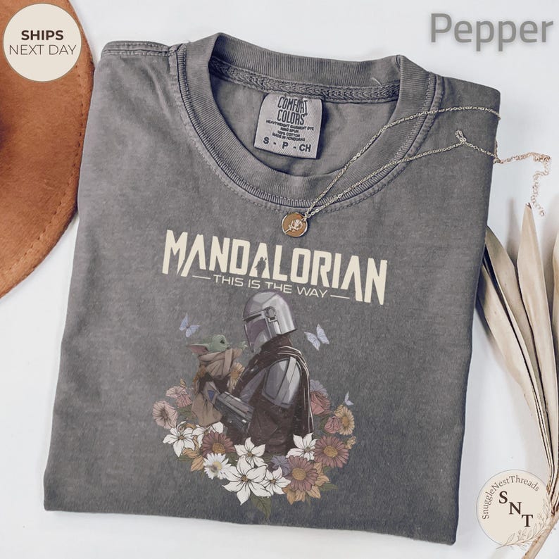 Puede incluir: Una camiseta de cuello redondo color pimienta con un gr&aacute;fico de The Mandalorian y Grogu rodeados de flores y mariposas. El texto dice "MANDALORIAN THIS IS THE WAY". Un collar de tono plateado con un peque&ntilde;o colgante descansa sobre la camiseta.