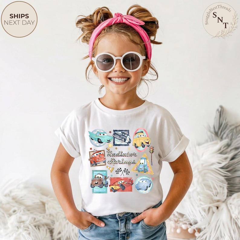 May include: A white t-shirt featuring a colorful graphic of various cartoon cars in stamp-like designs. The text "Radiator Springs" is printed in the center. The child is wearing sunglasses and a pink headband.