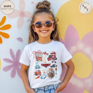 May include: A white t-shirt featuring colorful illustrations of characters from the movie "Cars". The design includes images of race cars, trucks, and other vehicles, with the word "Cars" in a red banner. The shirt is worn by a young person.