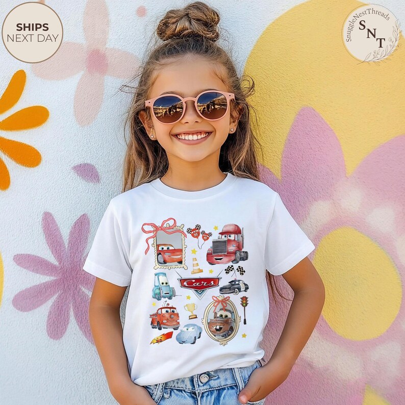 May include: A white t-shirt featuring colorful illustrations of characters from the movie "Cars". The design includes images of race cars, trucks, and other vehicles, with the word "Cars" in a red banner. The shirt is worn by a young person.