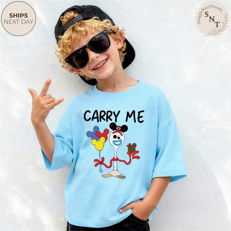 May include: A light blue t-shirt with the words "CARRY ME" in black. The shirt features a cartoon character holding balloons and a chocolate treat. The child is wearing sunglasses and a black cap.