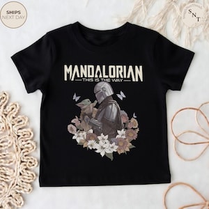 Puede incluir: Una camiseta negra de manga corta con un gr&aacute;fico del Mandaloriano y Grogu rodeados de flores y mariposas. El texto "MANDALORIAN" y "THIS IS THE WAY" est&aacute; impreso en una fuente de color crema sobre la ilustraci&oacute;n central de los personajes.