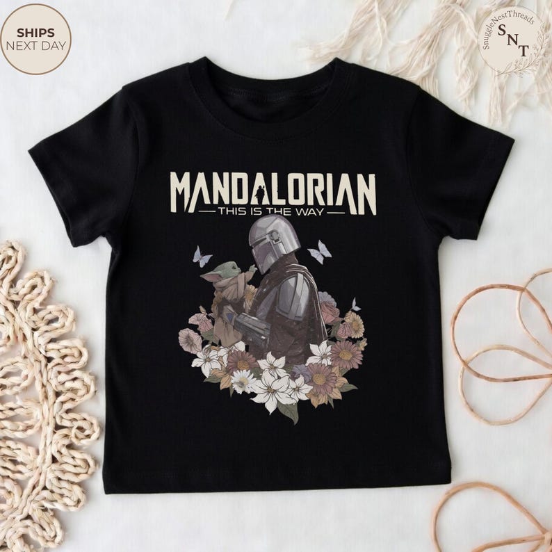 Puede incluir: Una camiseta negra de manga corta con un gr&aacute;fico del Mandaloriano y Grogu rodeados de flores y mariposas. El texto "MANDALORIAN" y "THIS IS THE WAY" est&aacute; impreso en una fuente de color crema sobre la ilustraci&oacute;n central de los personajes.