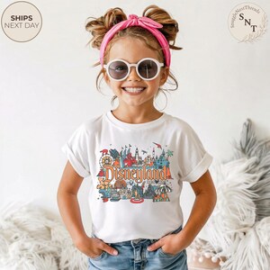 May include: A white t-shirt with a colorful Disneyland graphic. The graphic features various park icons and the word "Disneyland" in orange. The shirt is worn by a young person wearing sunglasses and a pink headband.
