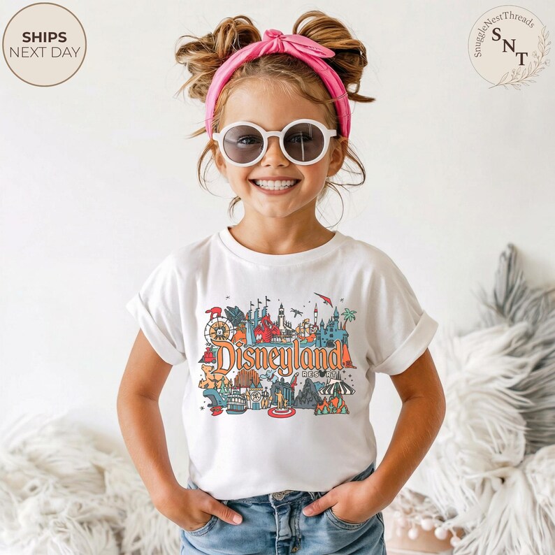 May include: A white t-shirt with a colorful Disneyland graphic. The graphic features various park icons and the word "Disneyland" in orange. The shirt is worn by a young person wearing sunglasses and a pink headband.