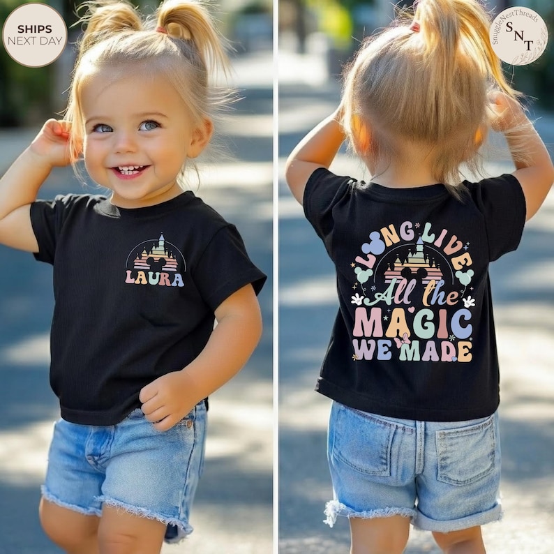 May include: Black t-shirt with a colorful castle graphic and the name "LAURA" on the front. The back of the shirt features the text "LONG LIVE All the MAGIC WE MADE" in pastel colors. The child is wearing denim shorts.