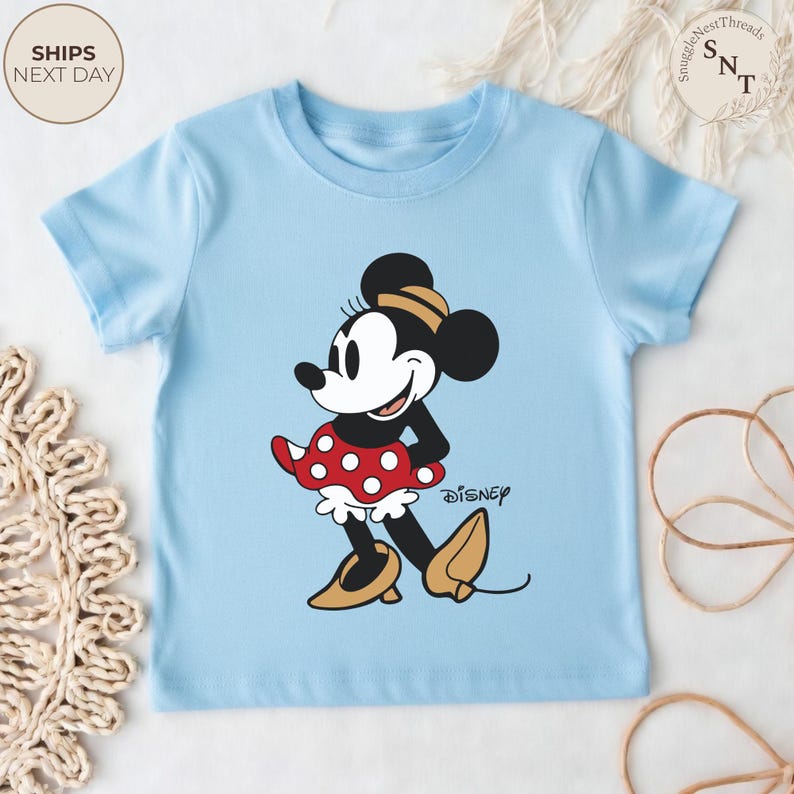 May include: A light blue t-shirt featuring a cartoon illustration of Minnie Mouse. Minnie Mouse is depicted in her classic outfit: a red polka-dot skirt, yellow shoes, and a black and yellow bow. The Disney logo is printed on the shirt.