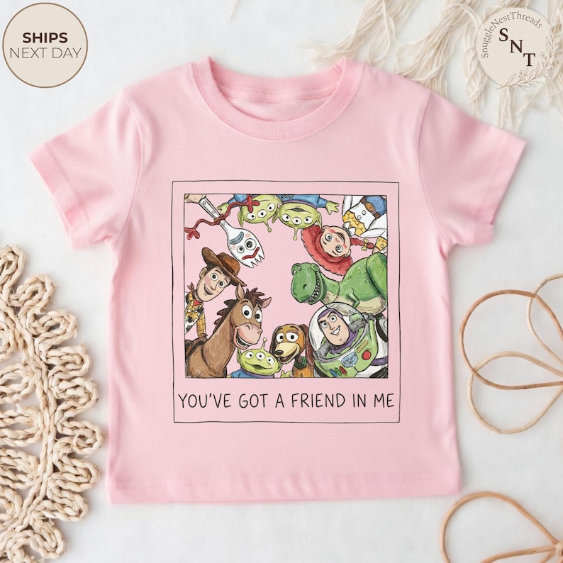 May include: A light pink t-shirt featuring a framed graphic of characters from the animated film "Toy Story." The graphic includes Woody, Buzz Lightyear, and other characters. The text "YOU'VE GOT A FRIEND IN ME" is below the graphic.