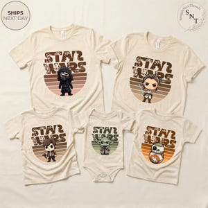May include: Beige Star Wars themed t-shirts and a onesie. Each shirt features a different character graphic with the words "STAR WARS" in a brown, retro-style font. The shirts have short sleeves and a crew neck.