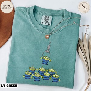 May include: A light green t-shirt featuring a graphic of the claw game with the little green aliens. The shirt has a crew neck and short sleeves. The text "LT GREEN" is at the bottom of the shirt.