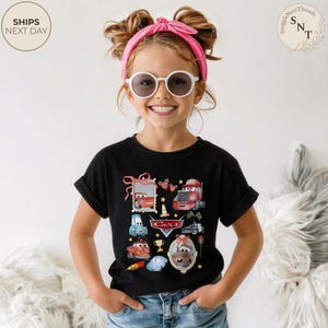 May include: Black t-shirt featuring a colorful Cars movie graphic. The design includes various car characters, a trophy, and the word "Cars" in a stylized font. The shirt is worn by a child.