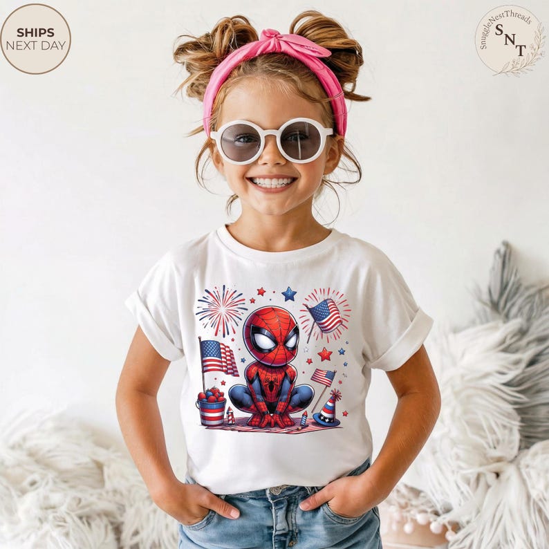 May include: A white short-sleeved t-shirt featuring a graphic of a cartoon Spider-Man character surrounded by American flags, fireworks, and a bucket of red, white, and blue items. The design includes small stars and festive patriotic elements.