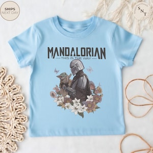 Puede incluir: Una camiseta de manga corta de color azul claro con un gr&aacute;fico del Mandaloriano y Grogu rodeados de flores y mariposas. El texto "MANDALORIAN" y "THIS IS THE WAY" est&aacute; impreso en negro sobre la ilustraci&oacute;n central de los personajes.