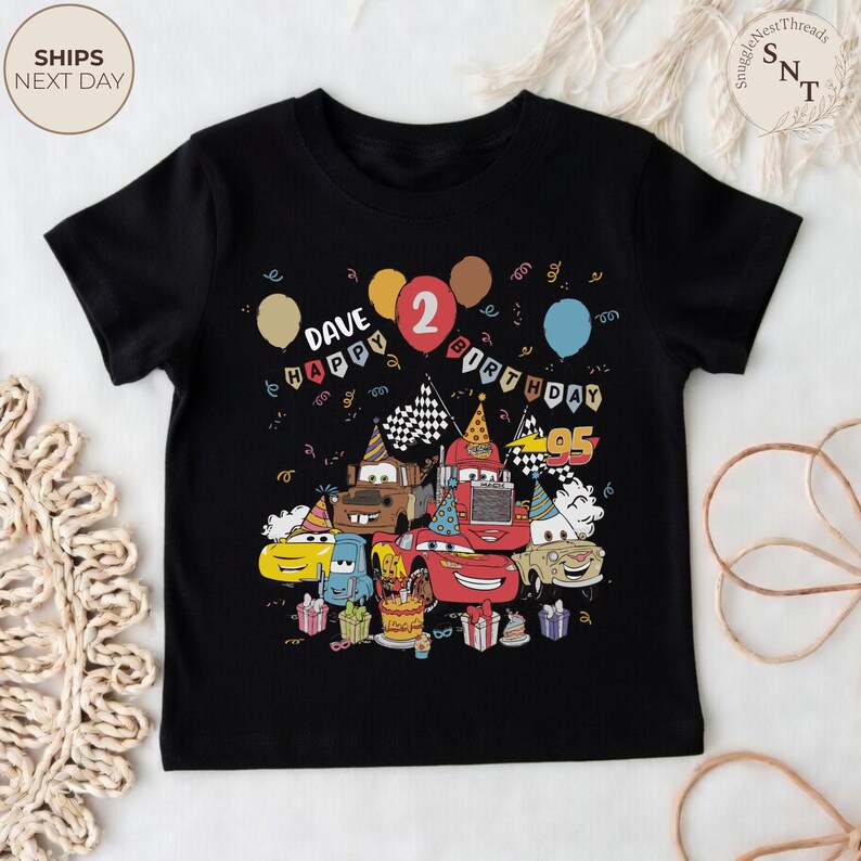 Puede incluir: Camiseta negra con un estampado colorido de coches de dibujos animados, globos y elementos de cumplea&ntilde;os. Incluye el texto "DAVE HAPPY BIRTHDAY 2". La imagen presenta una tarta de cumplea&ntilde;os y regalos, ideal para el segundo cumplea&ntilde;os de un ni&ntilde;o.