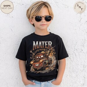 Puede incluir: Una camiseta negra con la palabra "MATER" y una imagen de dibujos animados de Mate, una gr&uacute;a marr&oacute;n de la pel&iacute;cula Cars. El gr&aacute;fico incluye efectos de rel&aacute;mpagos. El ni&ntilde;o lleva gafas de sol.