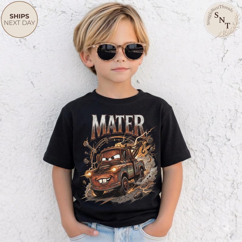 Puede incluir: Una camiseta negra con la palabra "MATER" y una imagen de dibujos animados de Mate, una gr&uacute;a marr&oacute;n de la pel&iacute;cula Cars. El gr&aacute;fico incluye efectos de rel&aacute;mpagos. El ni&ntilde;o lleva gafas de sol.
