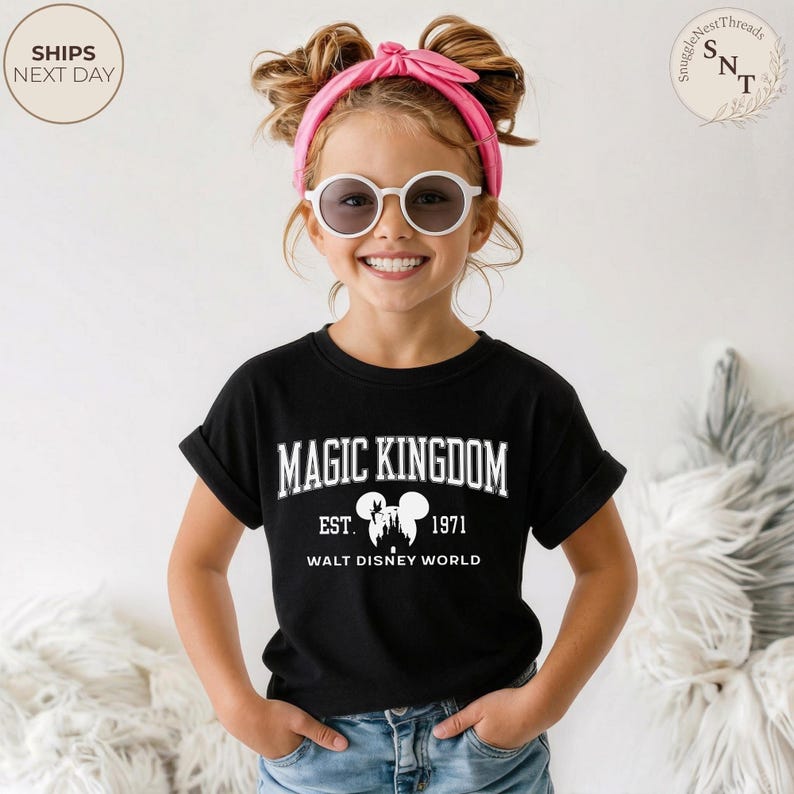May include: A black t-shirt with the words "MAGIC KINGDOM" in white block letters. Below the text is "EST. 1971 WALT DISNEY WORLD" with a silhouette of the Disney castle and Mickey Mouse ears. The shirt is worn by a child.