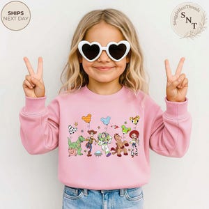 May include: A pink sweatshirt featuring a colorful graphic of Toy Story characters and balloons. The child is wearing white heart-shaped sunglasses and making a peace sign with their fingers. The text "SHIPS NEXT DAY" is in the upper left corner.