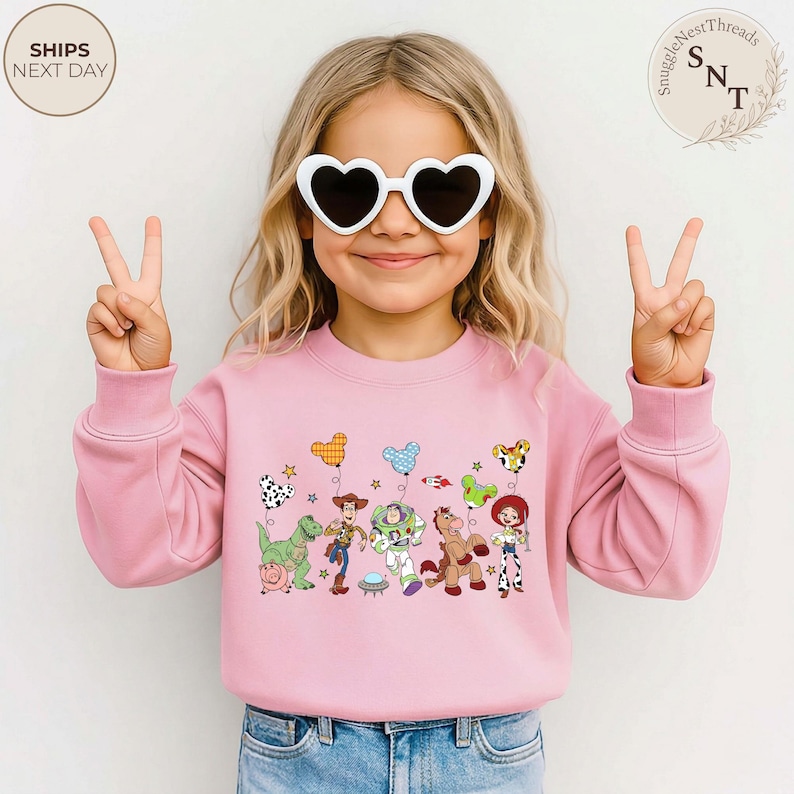 May include: A pink sweatshirt featuring a colorful graphic of Toy Story characters and balloons. The child is wearing white heart-shaped sunglasses and making a peace sign with their fingers. The text "SHIPS NEXT DAY" is in the upper left corner.