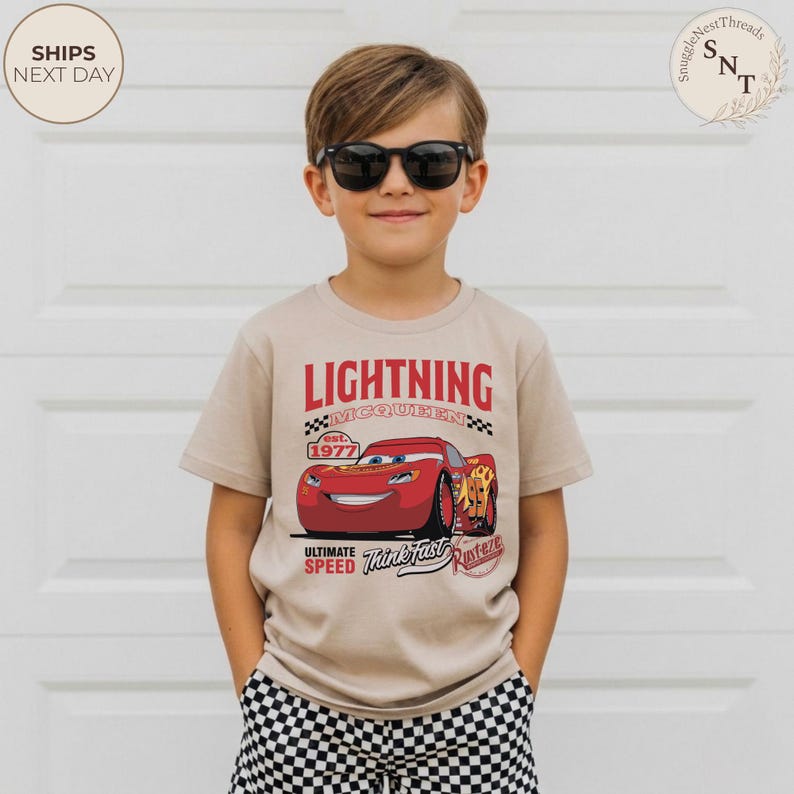 Op de afbeelding: Beige T-shirt met korte mouwen met een afbeelding van Lightning McQueen uit de film Cars. Het ontwerp bevat de tekst "LIGHTNING MCQUEEN est. 1977" en "ULTIMATE SPEED Think Fast Rusteze". De jongen draagt een zonnebril en een geruite korte broek.