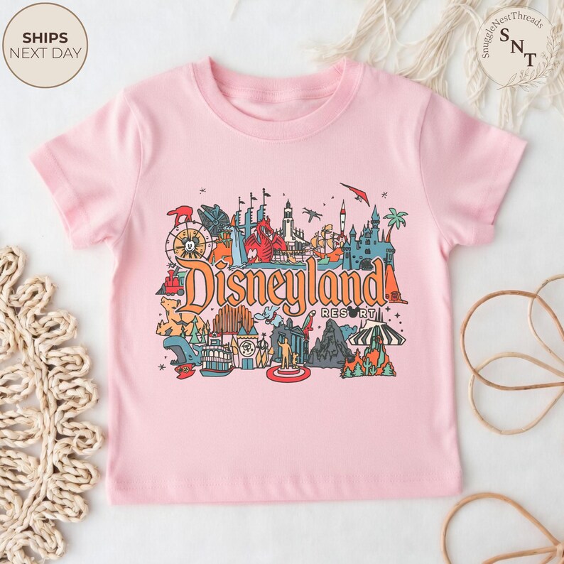 May include: A light pink t-shirt featuring a colorful graphic of Disneyland Resort. The graphic includes various park icons, the Disneyland Resort logo, and the text "Disneyland Resort". The shirt has short sleeves and a crew neck.