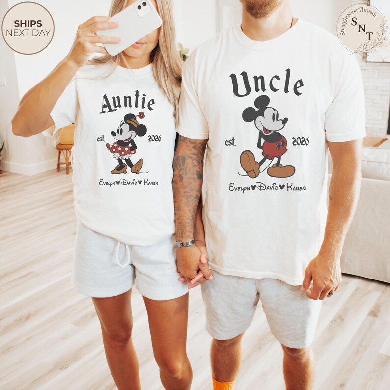 Pu&ograve; includere: Due magliette bianche sporche con grafica abbinata. Una dice "Auntie" con un design di Minnie Mouse, l'altra dice "Uncle" con un design di Mickey Mouse. Entrambe le magliette includono il testo "est. 2026" e un nome. Le magliette sono abbinate a pantaloncini grigi.