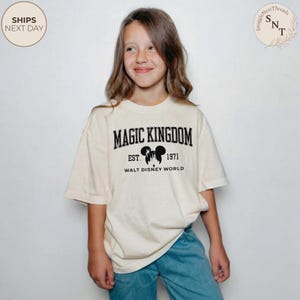May include: Cream-colored t-shirt featuring the text "MAGIC KINGDOM EST. 1971 WALT DISNEY WORLD" in black. The design includes a silhouette of Mickey Mouse ears. The shirt is worn by a person wearing blue jeans.