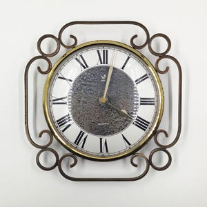 Può includere: Un orologio al quarzo rotondo con quadrante bianco, numeri romani neri e finiture color oro. L'orologio è racchiuso in una cornice decorativa in metallo marrone scuro con volute. Le lancette dell'orologio sono nere.