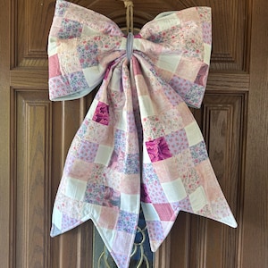 Op de afbeelding: Een grote decoratieve strik gemaakt van doorgestikte stof met een patchworkontwerp van diverse bloemen- en effen patronen in roze, lavendel en witte tinten. De strik is in het midden samengebonden met een lichtgrijs lint en heeft een lus van jute.