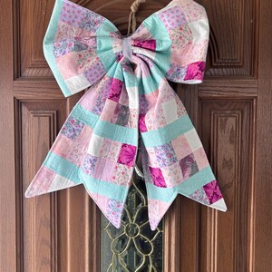 May include: A large, decorative bow made of patchwork fabric, featuring a mix of pink, purple, and teal squares. The bow is hanging on a wooden door. The bow is tied with a natural twine loop.