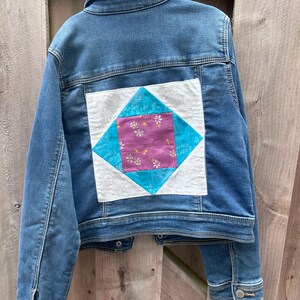 May include: A denim jacket with a quilted patch on the back. The patch is a square within a diamond shape, featuring a floral pattern in pink and white on a purple background, surrounded by turquoise and white fabric.